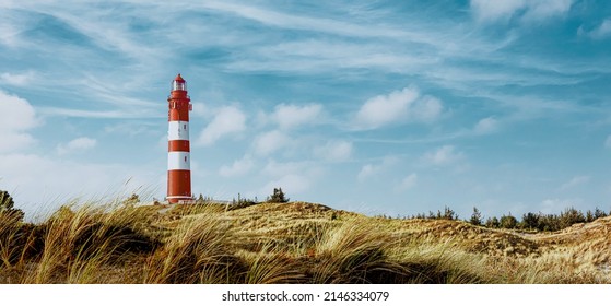 Tall red and white striped lighthouse on sand dunes in evening sunlight. Blue sky with white clouds. Summer vacation, holiday at sea concept.