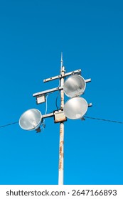A tall rusty pole with multiple large industrial floodlights and square LED lights mounted on metal brackets, set against a clear blue sky background.