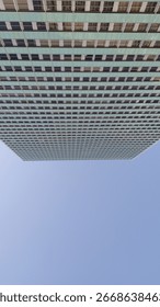 A tall skyscraper stretches vertically, emphasizing its modern facade against a clear blue sky. The repetitive grid pattern of the windows enhances the architectural symmetry, providing a sense of hei