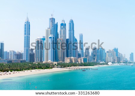 Tall skyscrapers of a modern, metropolitan cityscape tower over a beautiful, white, sandy beach on a warm, sunny day.