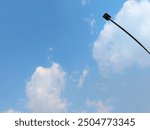 A tall, solitary streetlight set against a clear blue sky with scattered white clouds, capturing the simplicity of urban elements and the beauty of nature