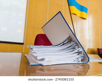 A tall stack of official documents in a blue file folder sits on a polished wooden table in a government office. The Ukrainian flag is visible in the background, alongside a projection screen and a re