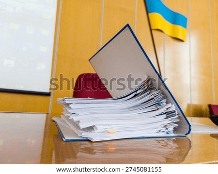 A tall stack of official documents in a blue file folder sits on a polished wooden table in a government office. The Ukrainian flag is visible in the background, alongside a projection screen and a re