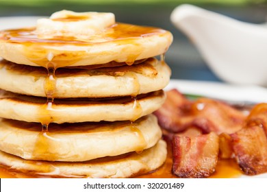 A tall stack of pancakes with butter and dripping syrup and three strips of bacon