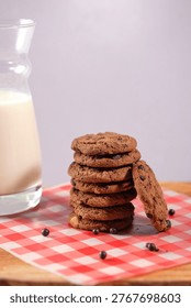 Tall stack of seven chocolate chip cookies with one leaning against the side, placed on a red and white gingham checkered cloth on a wooden surface. Glass milk carafe softly blurred on the left. Soft lavender background. Classic homestyle cookie stack concept.