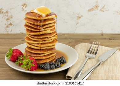 Tall Stack Of Warm Buttery Pancakes For Breakfast With Strawberries,  Blueberries And Syrup On White Plate, Wood Tabletop, Light Background, With Utensils And Napkin