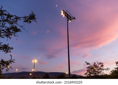 Tall Stadium Light Pole Shines Over A Baseball Field At Sunset With A Moon In The Sky