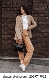 Tall stylish girl with a bag in a beige oversized jacket and orange pants on brick wall background - Shutterstock ID 2069749922