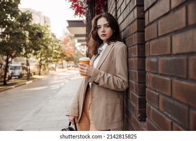 Tall stylish girl with coffee in a beige oversized jacket and orange pants on brick wall background - Shutterstock ID 2142815825