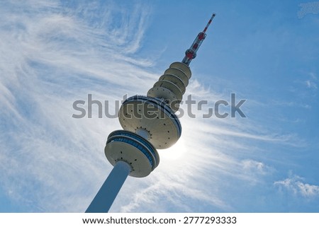 A tall telecommunications tower with multiple circular platforms and antennas stands against a bright blue sky with wispy clouds and sunlight behind it.