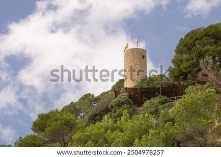 A tall tower with two flags on top of it. The sky is cloudy and the sun is shining. Castle Sant Joan, Lloret de Mar, Spain.