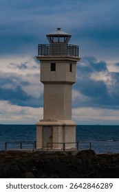 Tall white lighthouse standing on rocky shore against a dramatic cloudy sky and ocean horizon. A moody coastal scene showcasing maritime architecture and natural power