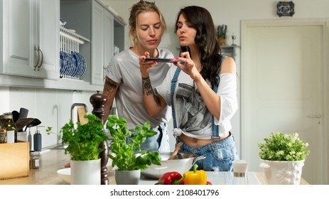 Tallinn, Harjumaa, Estonia - 06.17.2021: Isolated Beautiful Lesbian Couple Standing Behind The Kitchen Counter Cooking Food Together And Taking Photos Of The Food With A Smartphone.