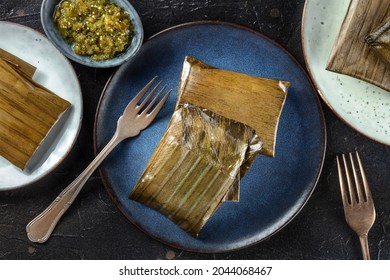 Tamal, Traditional Dish Of The Cuisine Of Mexico, Various Stuffings Wrapped In Green Leaves. Hispanic Food, Overhead Flat Lay Shot On A Black Background, With Salsa Verde