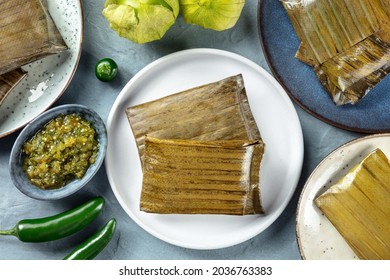 Tamales Oaxaquenos, Traditional Dish Of The Cuisine Of Mexico, Various Stuffings Wrapped In Green Leaves, Overhead Flat Lay Shot. Hispanic Food. With Chili Peppers, Tomatillos, And Salsa Verde