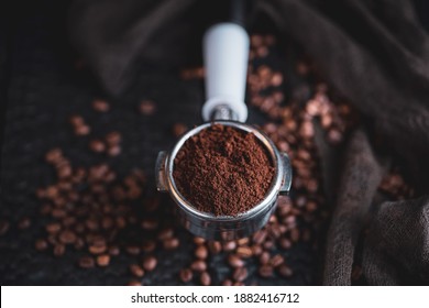 Tamped puck of coffee grounds within basket of portafilter and coffee beans spilled around in a dark and moody scene of natural light.