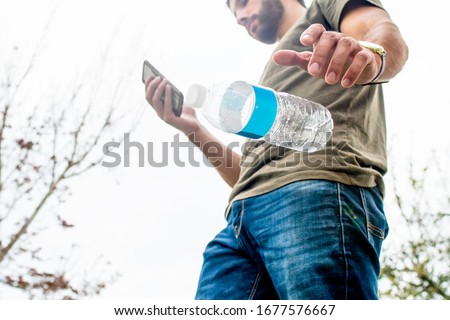A tan brown haired casual man walking through the park playing on his phone has no regard for the earth and its habitats as he drops his empty plastic water bottle to the ground paying no attention.