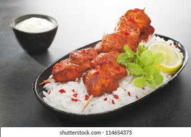 Tandoori chicken kebabs on a bed of rice, garnished with mint and lime, served with yoghurt, on a dark background. .More curry