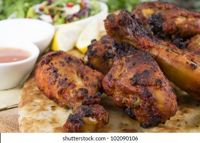 Tandoori chicken legs served on top of a naan bread, with salad, chili sauce, raita and lemon wedges.