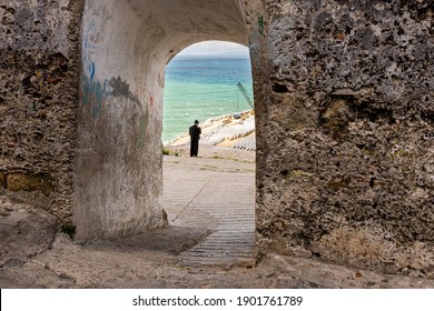 Tangier, Morocco - April 28, 2013: Old Man Wearing Beanie, Jacket And Trousers Looking Over Port Area, As Seen Through Archway Of Thick City Walls, Tangier, Morocco