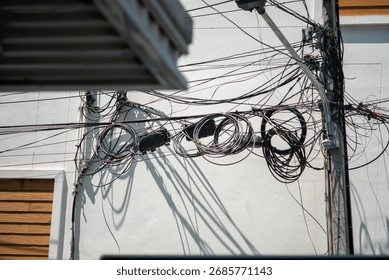 A tangled array of power lines casts intricate shadows against a building's white wall in an urban setting. The chaotic arrangement creates a striking visual contrast.
