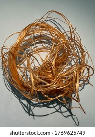 ​A tangled bundle of raw, fibrous material with a golden-brown or copper hue. The texture appears organic and stringy, resembling shredded plant fibers, unspun hemp, or coarse raffia. It is arranged in a loose, circular mess against a plain, light-colored background.
