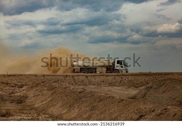 Tank truck and yellow dust cloud\
on sand road against grey cloudy sky. Road construction\
site.