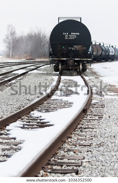 Tanker Train Cars with snow on a foggy winter\
day sitting on the far right set of tracks. Trees and buildings in\
the background. Vertical