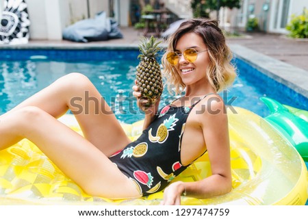 Tanned girl posing with interested smile while swimming in pool. Attractive short-haired woman relaxing at exotic resort and enjoying pineapple.