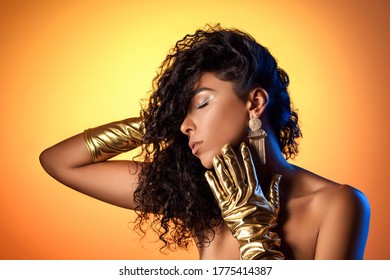 Tanned Luxurious Woman Posing In Golden Gloves, With Massive Earrings On A Gold Background. Vintage Retro Photo Shoot. Party Ideas