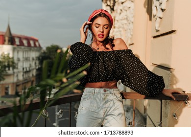 Tanned modern girl with curly hair and red lipstick in stylish wide long sleeve polka dot shirt and light jeans posing with closed eyes on balcony.. - Powered by Shutterstock