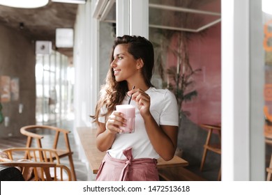 Tanned woman in bright outfit enjoys relaxing in cafe. Brunette drinking yogurt - Powered by Shutterstock