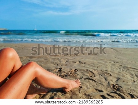 Tanned woman legs on the beach sand, sunbathing relaxation, seashore in the socond plan.