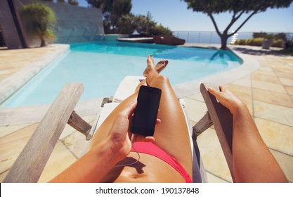 Tanned woman using smart phone by the poolside. Caucasian female model sunbathing on a deckchair and mobile phone. - Powered by Shutterstock