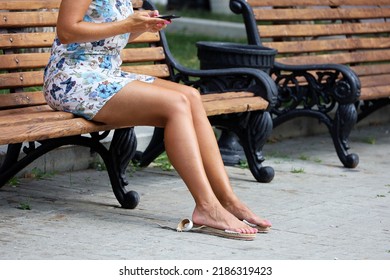 Tanned Woman With Wearing Short Summer Dress Sitting On Wooden Bench With Smartphone In Hand. Tired And Rubbed Legs With Removed Shoes, Sun Tan And Leisure In City Park