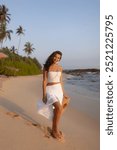 A tanned woman wearing a white skirt and top walks barefoot on a silent tropical beach at sunset, holding her sun hat. The serene setting with palm trees and calm waves makes it ideal for travel ads.