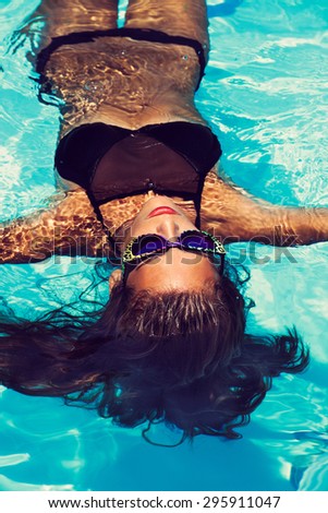 tanned young attractive woman in black bikini and sunglasses floating in pool