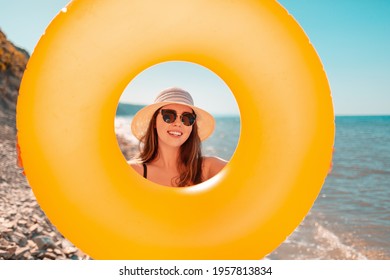 A Tanned Young Woman Holds An Orange Inflatable Circle And Looks Through It. In The Background Is A Wild Beach. Tint. The Concept Of A Summer Holiday On The Sea, Traveling And Swimming.