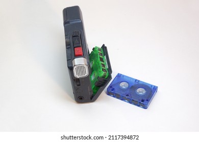 Tape Voice Recorder, Handheld, With Green Microcassette In Cover Of Tape Cassette Compartment. Red Button Of Rec Start, Silvery Electret Condenser Mic Housing, Blue Cassette Laying Near