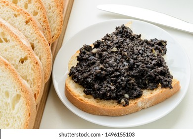 Tapenade Sandwich On A Sauser And Sliced Wheat Bread On A Wooden Chopping Board Over White Kitchen Table. Homemade Black Olive Paste With Anchovies, Garlic And Olive Oil. Top View.