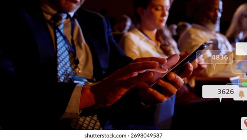Tapping man in dark suit checking smartphone in dim conference auditorium with notification bubbles. Corporate, networking, technology, professional, presentation, communication, modern