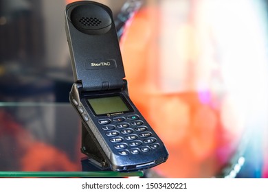 Taranto, Italy - 12 September 2019: Historic Motorola Mobile Phone Startac Model Photographed On A Crystal Table With A Graphic Background Of Orange And Gray Color.