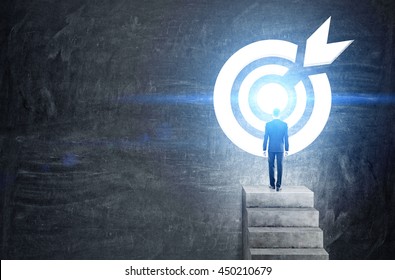Targeting concept with businessman on top of concrete ladder leading to abstract illuminated dartboard sketch on chalkboard background - Shutterstock ID 450210679