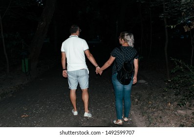 Targu Jiu, Romania. August, 30, 2019- A Man And A Blonde Woman Are Walking At Night In A Park Alley. A Couple Is Walking In The Park. Photographed From Behind