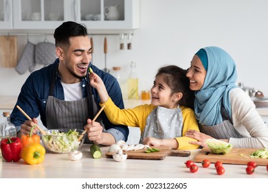 Taste It. Portrait Of Cute Little Arab Girl Feeding Her Daddy With Cucumber Slice, Happy Middle Eastern Family Of Three Cooking Healthy Tasty Food In Kitchen Together, Making Vegetable Meal At Home