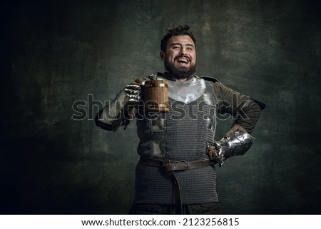 Tasting beer. Happy medieval warrior or knight with dirty wounded face holding big mug of beer isolated over dark vintage background. Comparison of eras, history, renaissance style, festival, ads