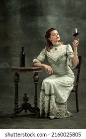 Tasting red wine. Conceptual portrait of young charming girl in image of medieval royal person or viscountess posing isolated on dark background. Comparison of eras concept, fashion, art, ad.