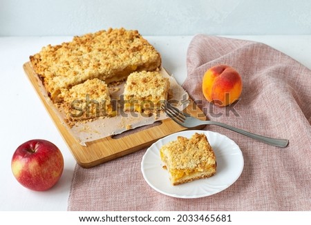 Tasty apple-peach shortcrust pie with crumble on wooden board and pink linen napkin.Piece of pie on saucer, fork, apple and peach. Homemade baking from seasonal fruits. Selective focus. Close up. 
