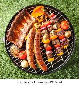 Tasty Assortment Of Meat On A Summer Barbecue With Sausages, Beef Kebabs And Spare Ribs With Tomatoes And Mushrooms, Overhead View Over Green Grass