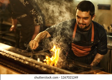 Tasty Beef Steak Cooked On Grill With Fire And Smoke. Beautiful Brutal Man With Bearded Face And Tattoo Cooking Meat. Professional Chef Wearing Apron In Restaurant Kitchen. 